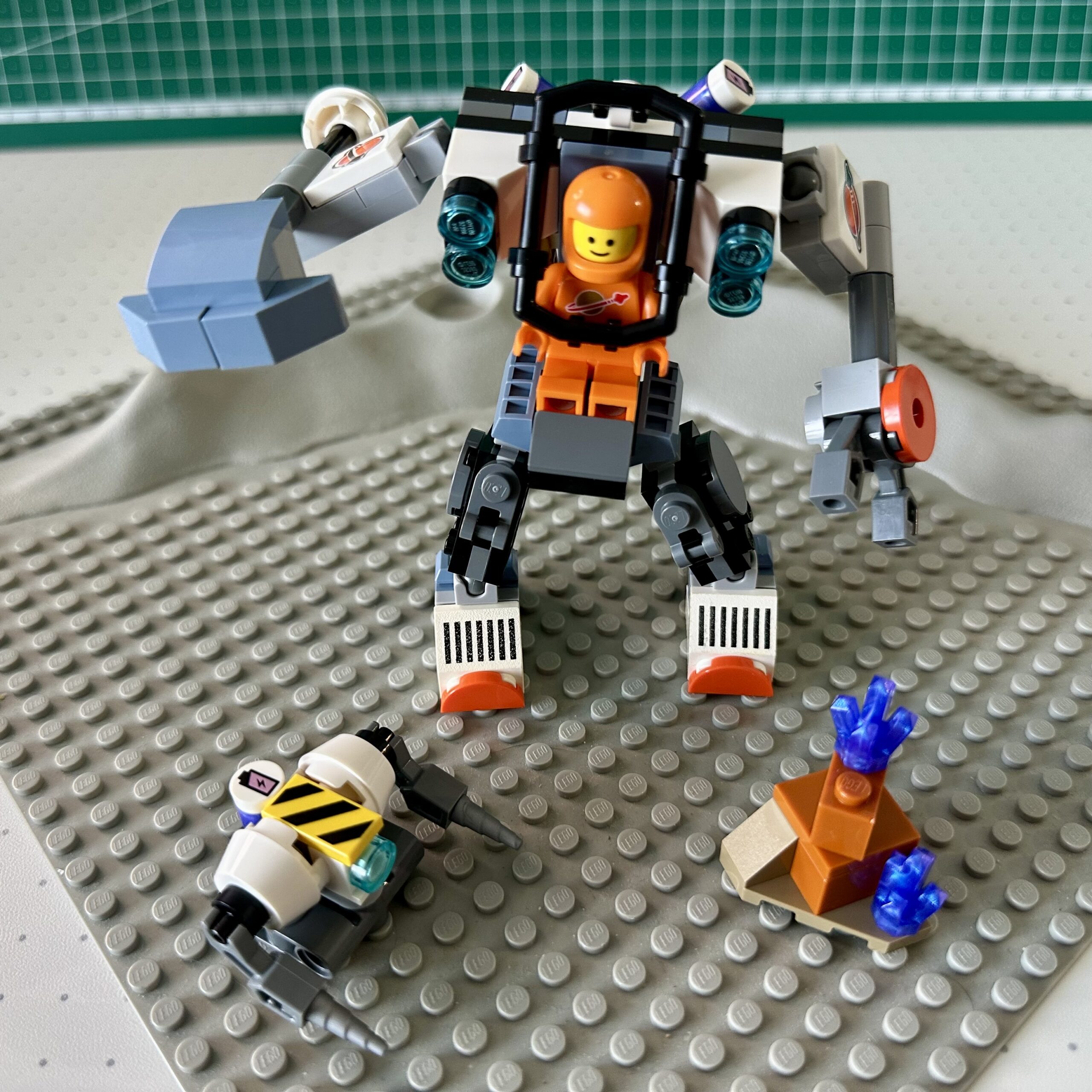 An orange LEGO classic spaceman drives the mech from the 2024 Space Construction Mech set. Posed on a classic gray crater baseplate from the 1970's. Some of the stickered parts have been replace with old printed elements from the 1980's.