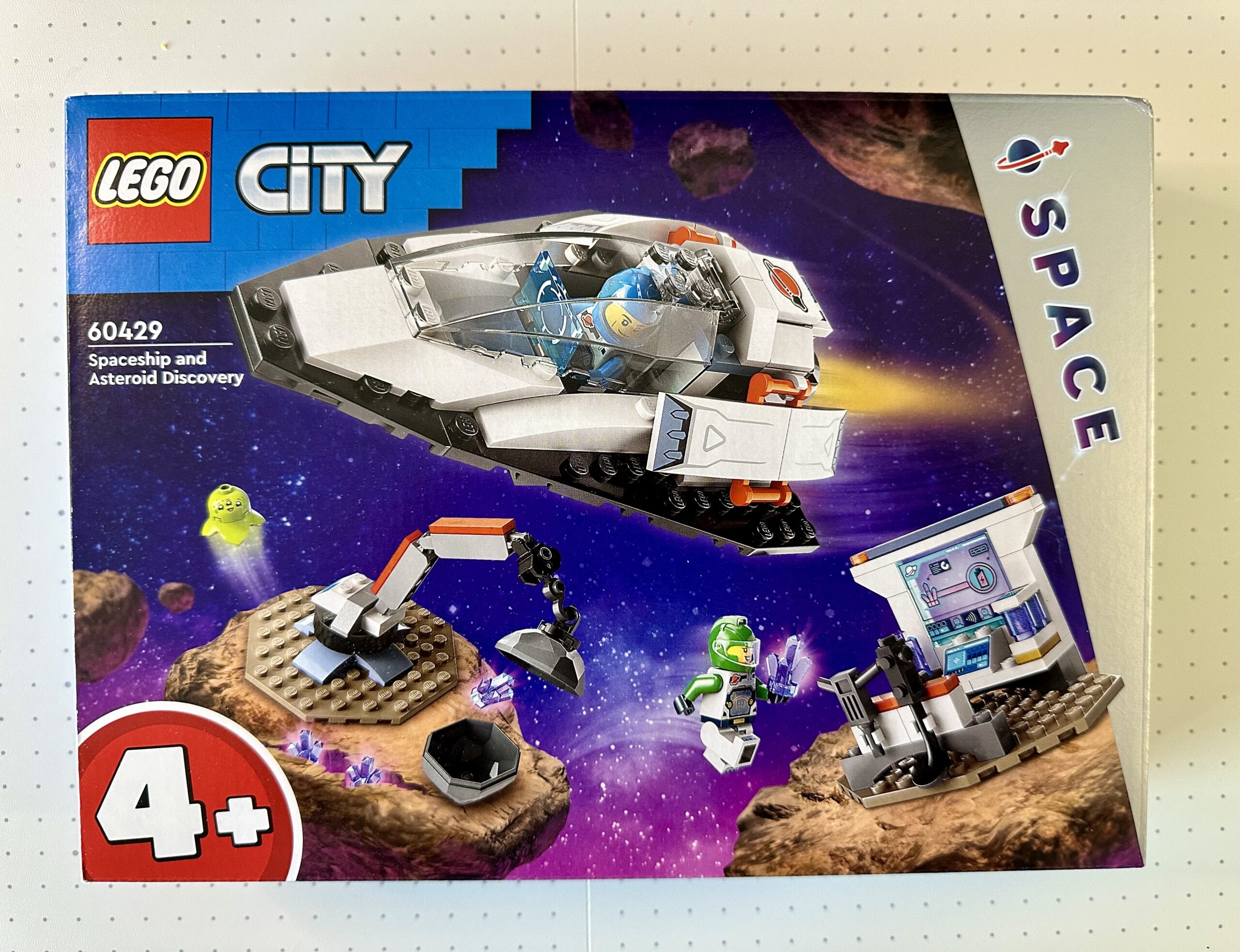 Box for LEGO City Space set 60429 Spaceship and Asteroid Discovery. Box depicts a spaceship flying over a couple pieces of alien terrain; one with a crane opening a boulder and another with a research station.